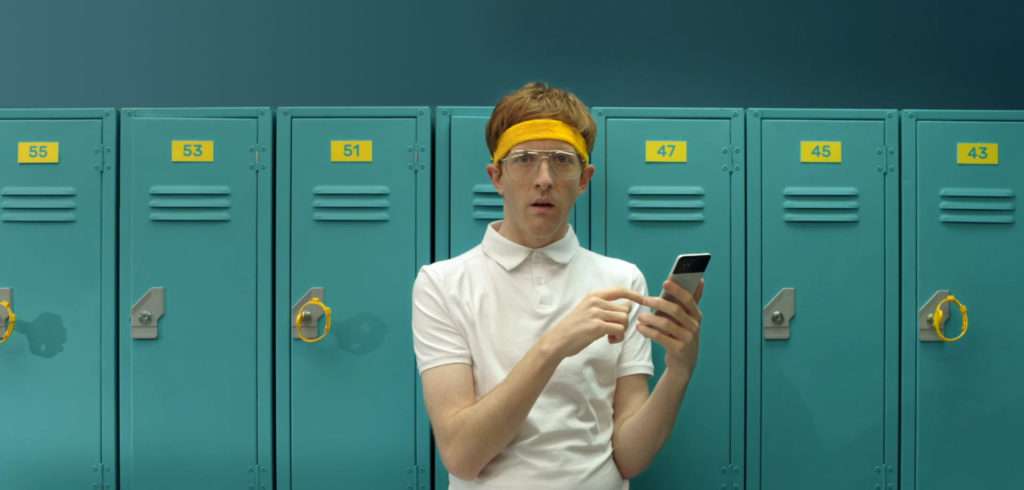 a young man with ginger hair has a yellow band in his hair, wearing a white polo-shirt holding a smart phone and in the background blue lockers with yellow numer-tags