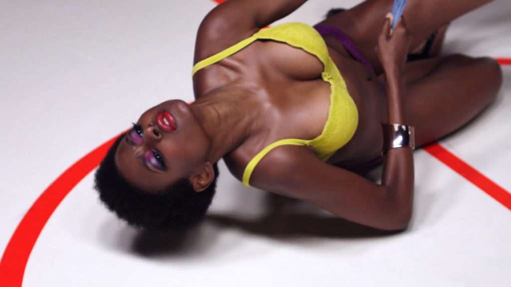 a coloured woman is lying on the floor, she wears a lot of make-up, a yellow bra and there are red lines on the floor