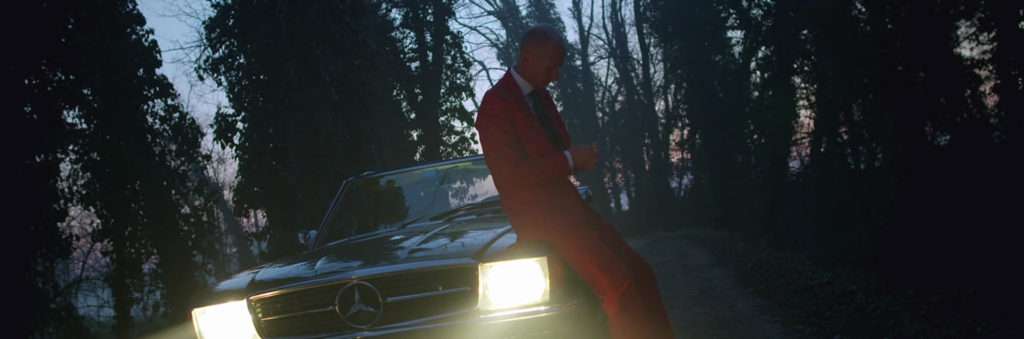 a man in a red suit is sitting on the front of an old Mercedes car, the lights are on and night is falling